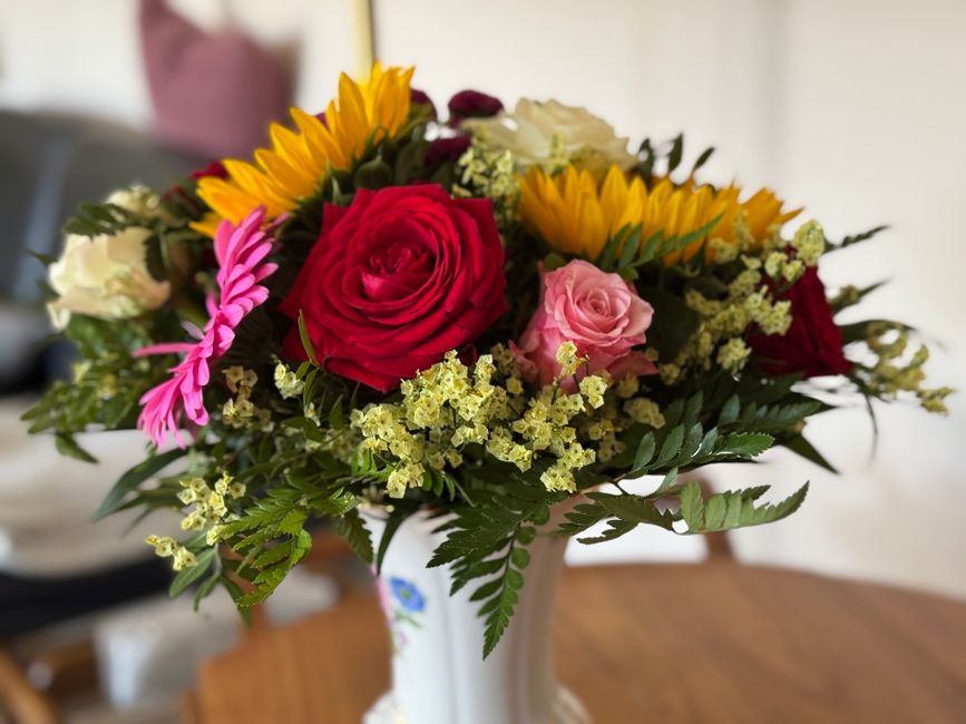 A vibrant floral bouquet arranged in a white ceramic vase, featuring large red roses, soft pink roses, bright yellow sunflowers, and sprigs of small white filler flowers, complemented by green fern le