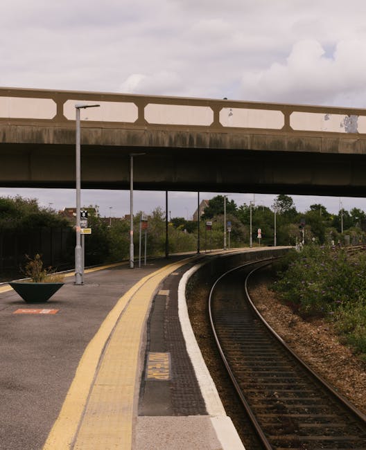 A view of a quiet train platform with a curved railway track running alongside it, featuring a yellow safety line and a textured tactile strip at the edge. The platform is partially shaded by an overp
