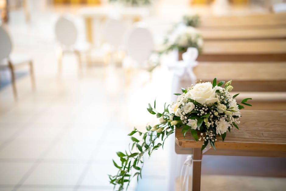 A bouquet arrangement of fresh white flowers, including roses and baby's breath, with green foliage, resting on the wooden pew of an indoor chapel or wedding venue. The bouquet appears to be handmade,