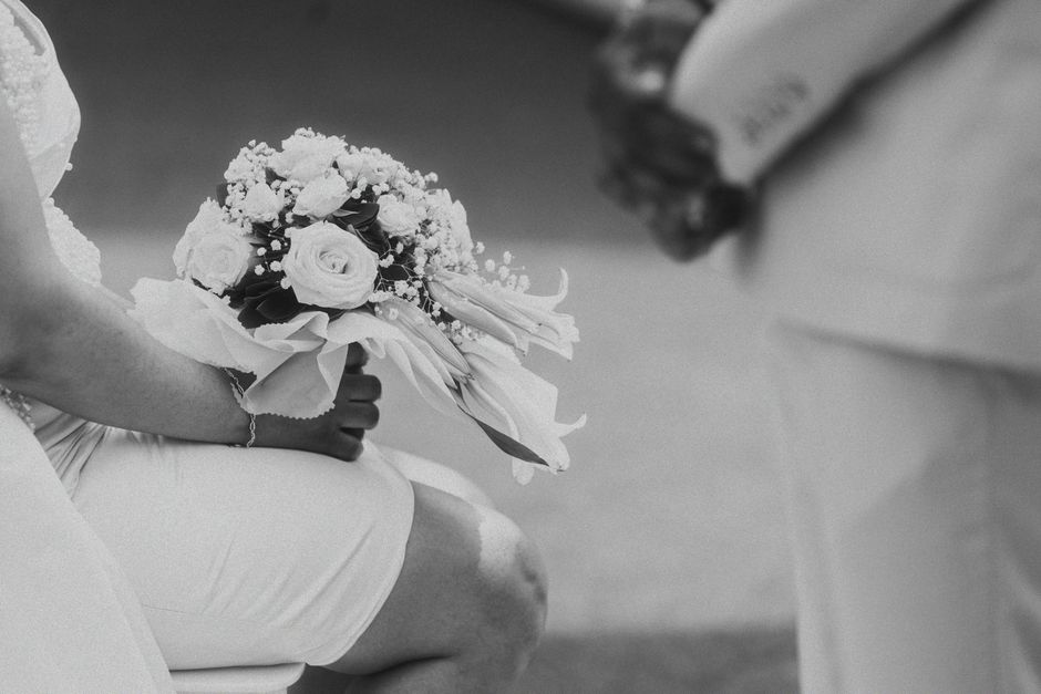 A bride seated, holding a floral bouquet featuring roses, baby's breath, and other delicate blooms in soft, pastel shades, wrapped in elegant tissue paper and a satin ribbon. The bouquet showcases fre