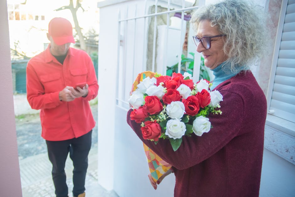 A mature woman with curly gray hair and wearing glasses, dressed in a maroon sweater and light blue turtleneck, stands near a white door holding a vibrant floral gift bouquet consisting of red and whi
