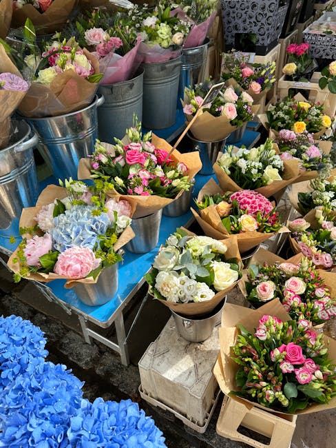 A display of various fresh floral bouquets arranged in metallic and paper-wrapped containers at a flower shop. The bouquets feature pink, white, and blue flowers, including roses, hydrangeas, and othe