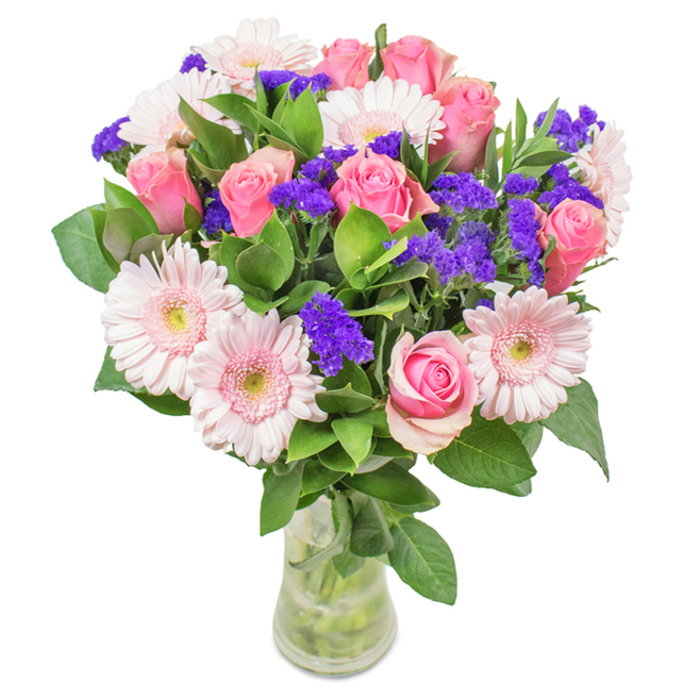 A captivating floral arrangement from Florists Catford sits in a simple glass vase, displayed against a pristine white background. Seven to nine lush pink roses, in varying stages of bloom from tightly furled buds to expanding spirals, form the radiant core. Around them, five to seven pale pink gerbera daisies showcase their large, cheerful faces-each petal softly arching out from nearly white centers that catch gentle light, reminiscent of Catford Bridge's morning ambience. Intertwined throughout are lively sprigs of vibrant purple statice, their texture and colour echoing the artistic verve of Catford's community. The bouquet is anchored by dense, deep green foliage that adds volume and a whisper of woodland fragrance, conjuring the green pockets along Ravensbourne Park. Every visible stem in the crystal-clear vase testifies to the bouquet's freshness and the artisanal touch of Catford's local florists. This arrangement, with its rosy warmth and uplifting palette, is suited for birthdays, anniversaries, or to brighten a loved one's day across Catford-gifted with a sense of affection, celebration, and place.